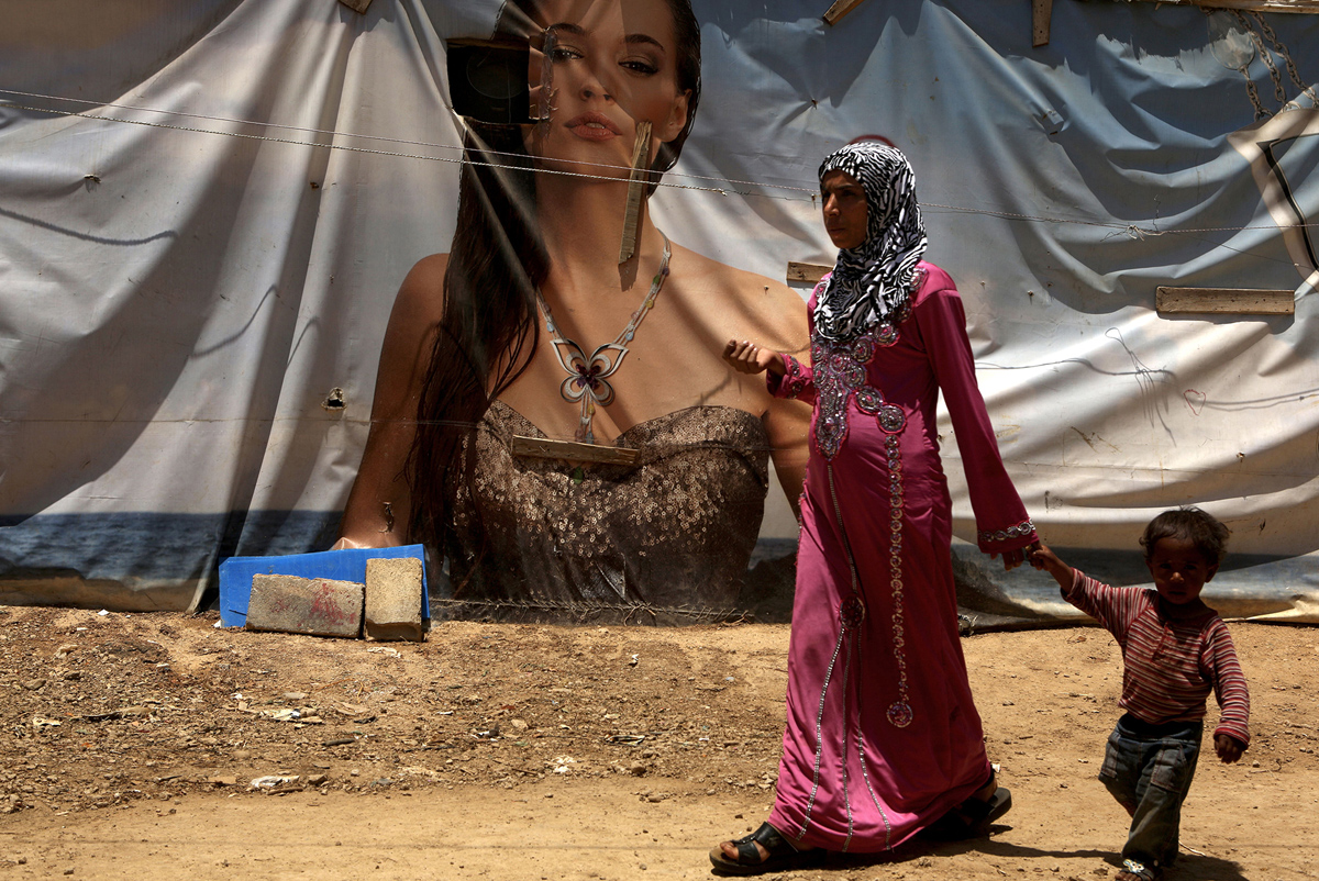 A Syrian refugee from Aleppo holding her son's hand walks past an advertising banner used as a makeshift tent in a Syrian camp
